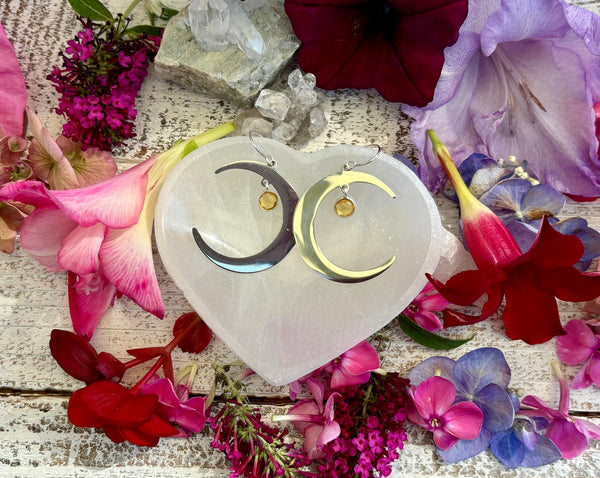 Amazing Earth Half Moon Earrings with sterling silver citrine drops on sterling silver backs, displayed on a heart-shaped crystal and surrounded by vibrant pink, purple, and red flowers on a rustic white surface.