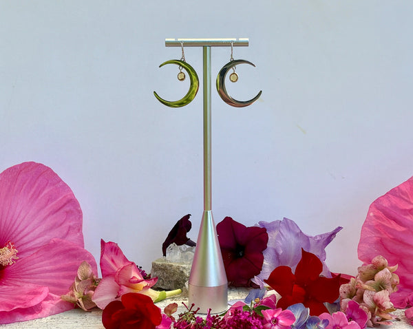 Half Moon Earrings with Sterling Silver Citrine Drops by Amazing Earth hang on a silver stand, surrounded by vibrant pink, red, and purple flowers against a light background.