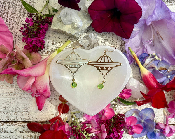 Amazing Earth’s Planet Earrings with Gold Peridot on 14KG backs rest on a white heart-shaped dish, surrounded by colorful flowers and crystals atop a rustic white wooden surface.