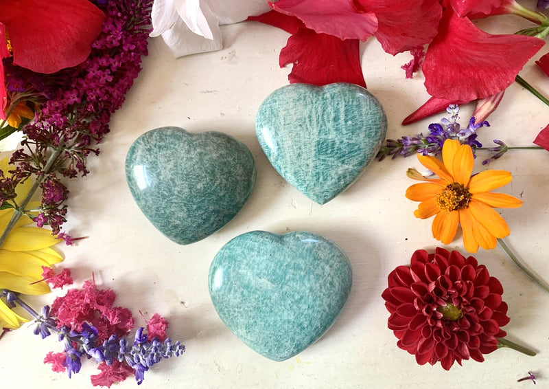 three green crystal hearts on tabletop with flowers in background.