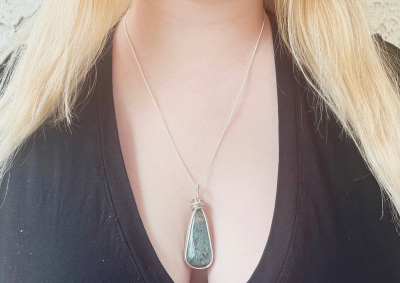 Tear drop shaped moss agate sterling wrapped pendant hanging on sterling chain around model's neck. 