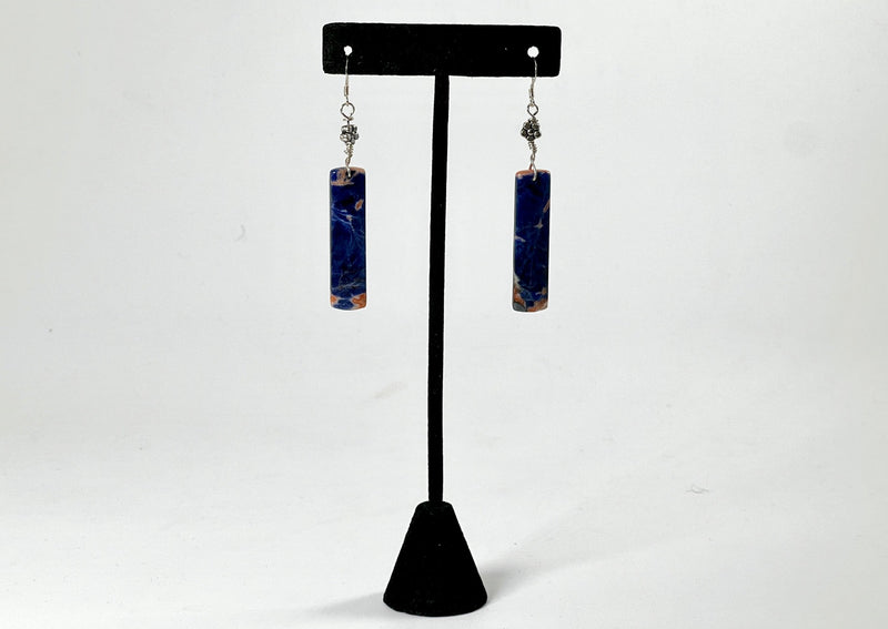 Rectangular dangling blue orange sodalite earrings on silver wire with flower detail hanging from black t stand. 